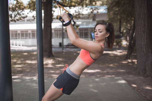 Pretty young girl doing sports on the playground with TRX straps. Healthy lifestyle concept, training on trx. Total Body Resistance Exercises. Summer time. - Foto, Imagen