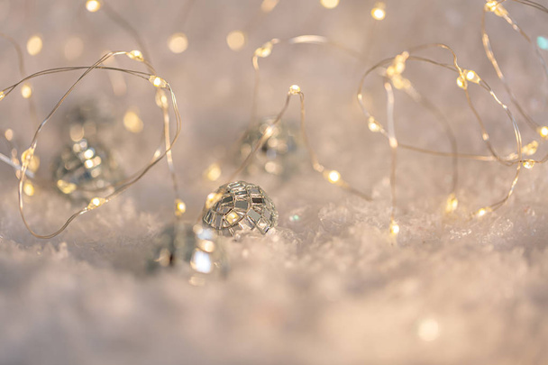Small decorative balls with a mirror and a luminous garland on a snow. Blurred festive gray background with white bokeh. - Photo, image