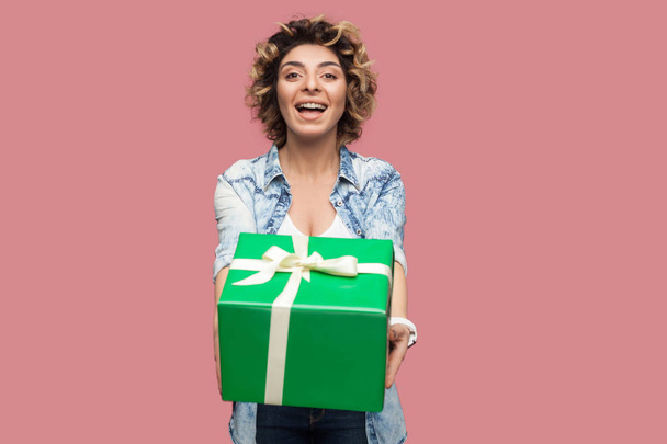 happy beautiful young woman in blue shirt with curly hairstyle standing and giving green gift box with toothy smile while looking at camera on pink background - Foto, imagen