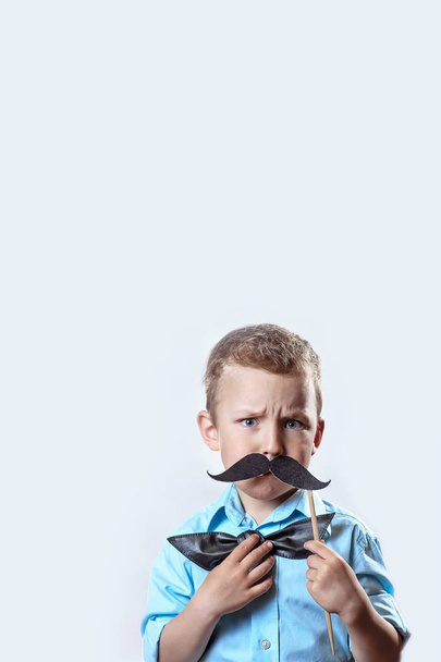 frowning serious boy in a light shirt put a mustache on a stick and a bow tie on his face to make him look older. - Φωτογραφία, εικόνα