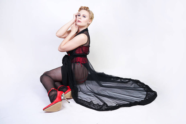 beautiful chic plus size blonde with short hair in red lace corset and black sheer long robe, in sexy pantyhose with seam and red patent leather shoes with steel heel posing on white background alone - Φωτογραφία, εικόνα