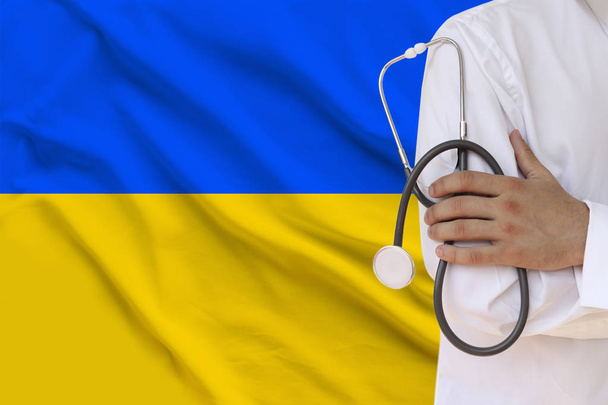 male doctor in white coat holding in his hand a medical instrument with a stethoscope against the background of the national flag, close-up, concept, copy space - Photo, image