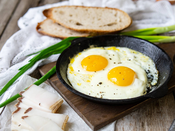 Scrambled eggs in frying pan with pork lard, bread and green feathers onions on old wooden table. National Ukrainian or belorussian food. Breakfast, lunch. Close up, selective focus, rustic style. - 写真・画像