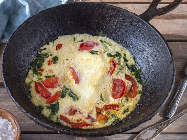 Omelet with tomatoes in a round cast iron pan on an old Board table, cotton napkins and coarse salt in a clay salt shaker, table knife, taken at close range - Photo, Image