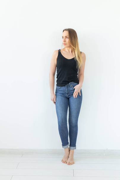 Styl a lidi koncept-mladá žena v džínách, která stojí nad bílou stěnou a vypadá jako sexy - Fotografie, Obrázek