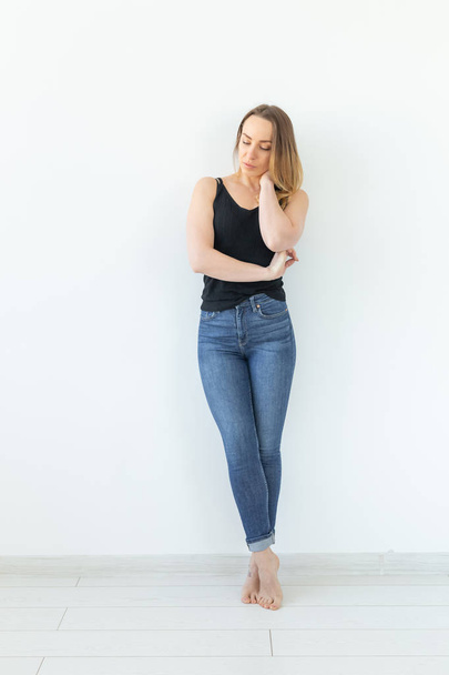 Estilo y concepto de la gente - mujer joven en jeans de pie sobre la pared blanca y se ve como sexy
 - Foto, Imagen