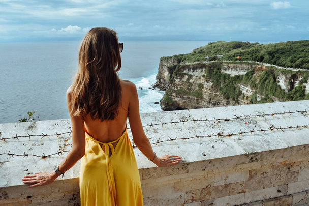 Asia travel series. Beautiful woman with dark hair in elegant yellow dress admiring a panoramic view of the ocean - Fotó, kép