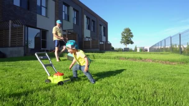 Людина косіння газон з газонокосарка в той час як син грає з іграшкою газонокосарка. Карданний - Кадри, відео