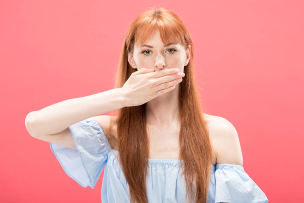 front view of serious redhead girl covering mouth with hand and looking at camera isolated on pink - Foto, Imagem