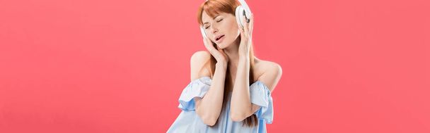 plano panorámico de atractiva pelirroja escuchando música en auriculares con los ojos cerrados aislados en rosa
 - Foto, Imagen