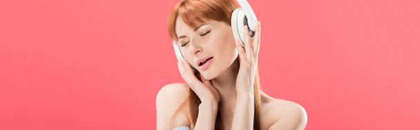 plano panorámico de atractiva pelirroja escuchando música en auriculares con los ojos cerrados aislados en rosa
 - Foto, Imagen