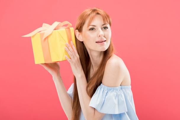 curious redhead birthday girl holding yellow gift box with ribbon isolated on pink - Photo, image