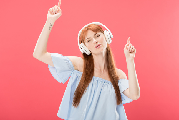 mulher ruiva atraente ouvindo música em fones de ouvido com olhos fechados e dançando isolado em rosa
 - Foto, Imagem