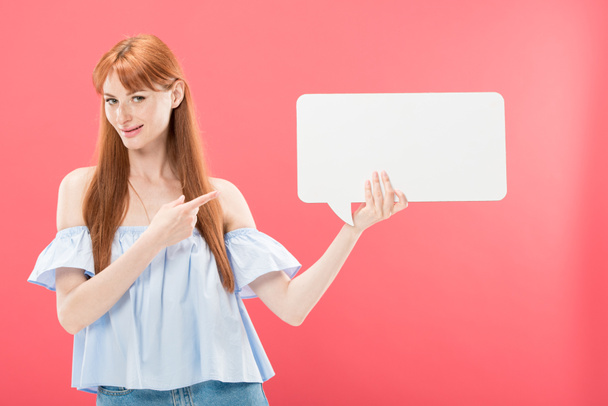 redhead young woman pointing with finger at empty speech bubble and looking at camera isolated on pink - Photo, Image