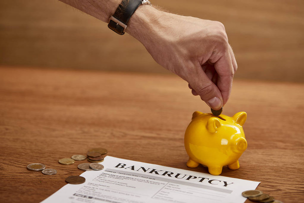vista cortada do homem colocando moeda no banco piggy amarelo perto da forma de falência
 - Foto, Imagem