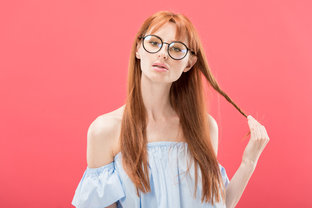 front view of redhead girl in glasses and stylish blouse playing with hair and looking at camera isolated on pink - Foto, immagini