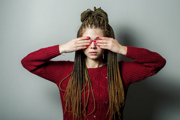Close up shot of young pretty caucasian teenage girl with african braids and problem skin and in maroon sweater covering her eyes with her hands. - Foto, imagen