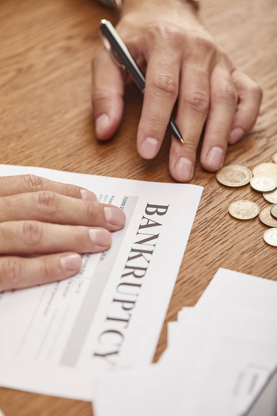 partial view of businessman filling in bankruptcy form at wooden table with coins - Photo, Image