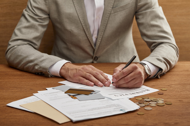 vista parcial del hombre de negocios en traje llenando formulario de bancarrota en la mesa de madera con documentos y tarjetas de crédito
 - Foto, Imagen