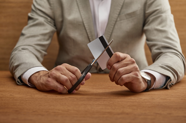 cropped view of businessman in suit cutting credit card with scissors at wooden table  - Foto, Bild