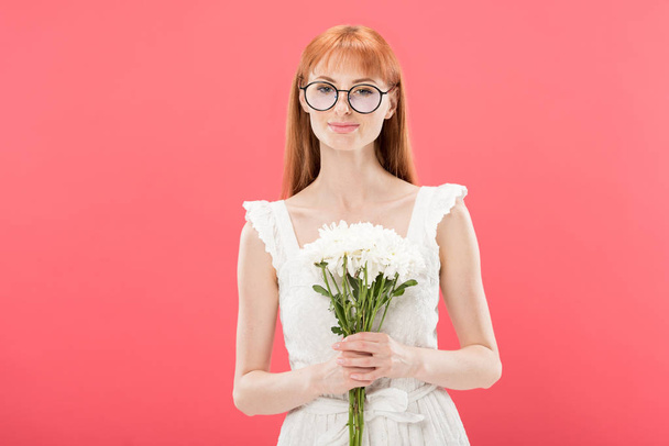 front view of attractive redhead girl in glasses and white dress holding flowers and smiling isolated on pink - Photo, Image