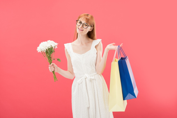 smiling redhead girl in glasses and white dress holding bouquet of flowers and shopping bags isolated on pink - Foto, Imagen