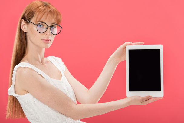 gyönyörű vöröshajú lány a szemüveg és fehér ruhában gazdaság digitális tabletta, üres képernyő és néztem kamera izolált rózsaszín - Fotó, kép