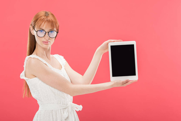 hermosa pelirroja en gafas y vestido blanco sosteniendo tableta digital con pantalla en blanco y mirando a la cámara aislada en rosa
 - Foto, Imagen