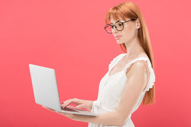 serious redhead young woman in glasses using laptop isolated on pink - Zdjęcie, obraz