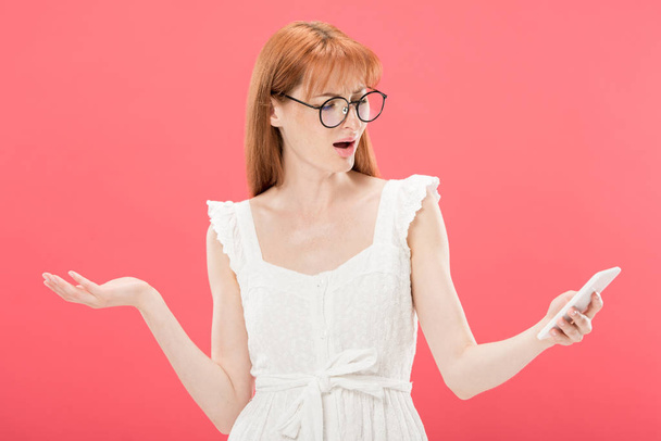 niezadowolony rudowłosy Kobieta w okularach i białej sukni za pomocą smartfona izolowane na różowy - Zdjęcie, obraz
