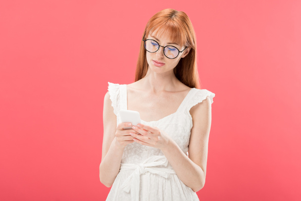 redhead woman in glasses and white dress using smartphone isolated on pink - Photo, image
