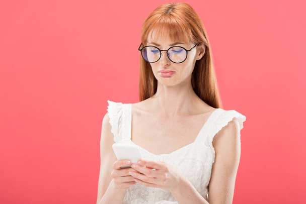 displeased redhead young woman in glasses and white dress using smartphone isolated on pink - Photo, Image