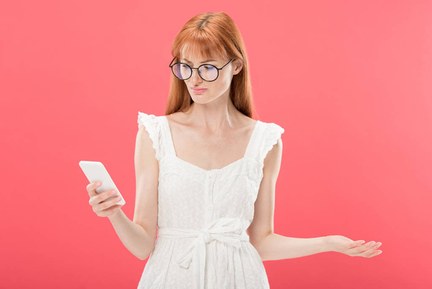 displeased redhead young woman in glasses and white dress using smartphone isolated on pink - Valokuva, kuva
