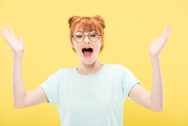 pleased redhead girl in glasses and t-shirt gesturing isolated on yellow - Fotó, kép