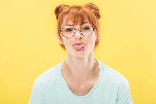 front view of cheerful redhead girl in glasses sticking out tongue and looking at camera isolated on yellow - Zdjęcie, obraz