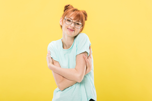 ευχαριστημένος κοκκινομάλλα κορίτσι με τα γυαλιά και το μπλουζάκι αγκαλιάζοντας τον εαυτό της και κοιτάζοντας την κάμερα με χαμόγελο απομονώνεται σε κίτρινο - Φωτογραφία, εικόνα