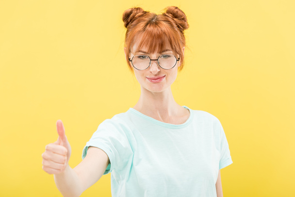 front view of smiling redhead girl in glasses and t-shirt looking at camera and showing thumb up isolated on yellow - Photo, Image