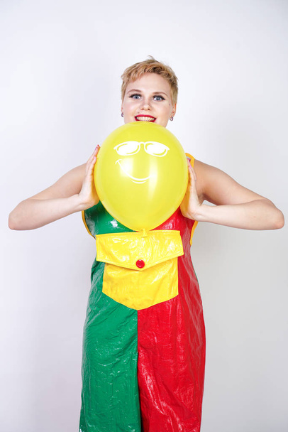 cute chubby girl with short blonde hair and plus size figure in bright colored PVC jumpsuit posing with air balloons on white background alone. playful young woman in halloween good clown costume. - Photo, Image