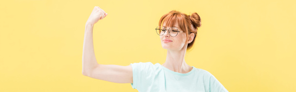 panoramic shot of confident pleased redhead girl in glasses and t-shirt holding fist up isolated on yellow - Photo, Image