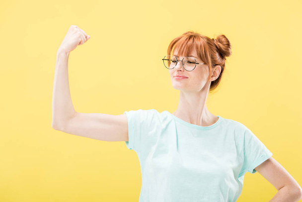 confident pleased redhead girl in glasses and t-shirt holding fist up isolated on yellow - Φωτογραφία, εικόνα