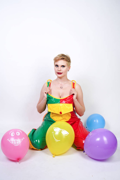 cute chubby girl with short blonde hair and plus size figure in bright colored PVC jumpsuit posing with air balloons on white background alone. playful young woman in halloween good clown costume. - 写真・画像