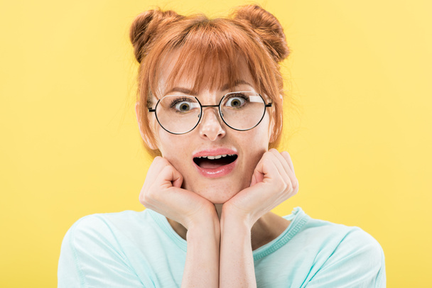 front view of surprised redhead girl in glasses propping face with hands and looking at camera isolated on yellow - Fotoğraf, Görsel