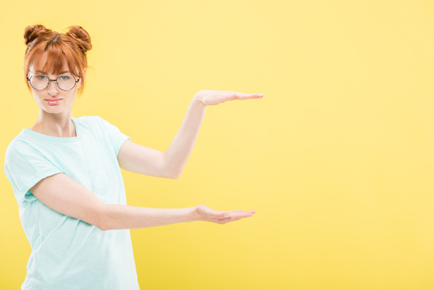 attractive redhead girl in t-shirt and glasses pointing with hands and looking at camera isolated on yellow - Foto, immagini