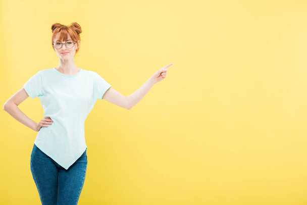 smiling redhead girl in glasses standing with hand on hip and pointing with finger isolated on yellow - Fotoğraf, Görsel