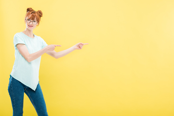 smiling redhead girl in glasses and t-shirt pointing with fingers and looking at camera isolated on yellow - Photo, Image