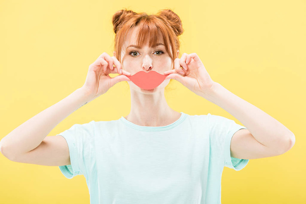 front view of redhead girl in t-shirt holding paper lips and looking at camera isolated on yellow - Foto, Bild