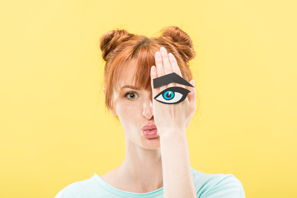 front view of redhead girl holding paper eye and eyebrow and looking at camera isolated on yellow - Photo, Image