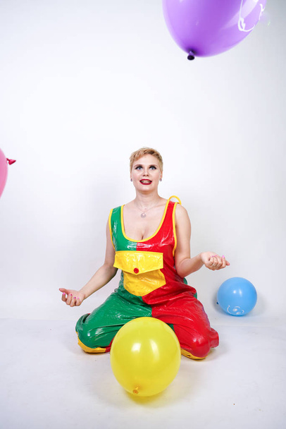 cute chubby girl with short blonde hair and plus size figure in bright colored PVC jumpsuit posing with air balloons on white background alone. playful young woman in halloween good clown costume. - Valokuva, kuva