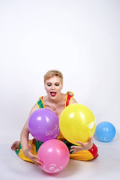 cute chubby girl with short blonde hair and plus size figure in bright colored PVC jumpsuit posing with air balloons on white background alone. playful young woman in halloween good clown costume. - Foto, Imagen