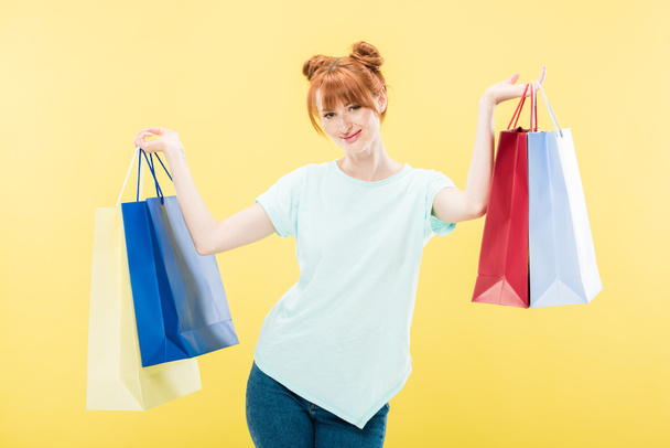 smiling redhead girl holding shopping bags and looking at camera isolated on yellow - Фото, изображение